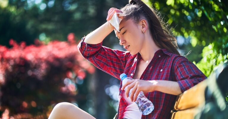 Tired sweating woman wipes her forehead with a napkin and holds cold water bottle in a park in summer hot weather