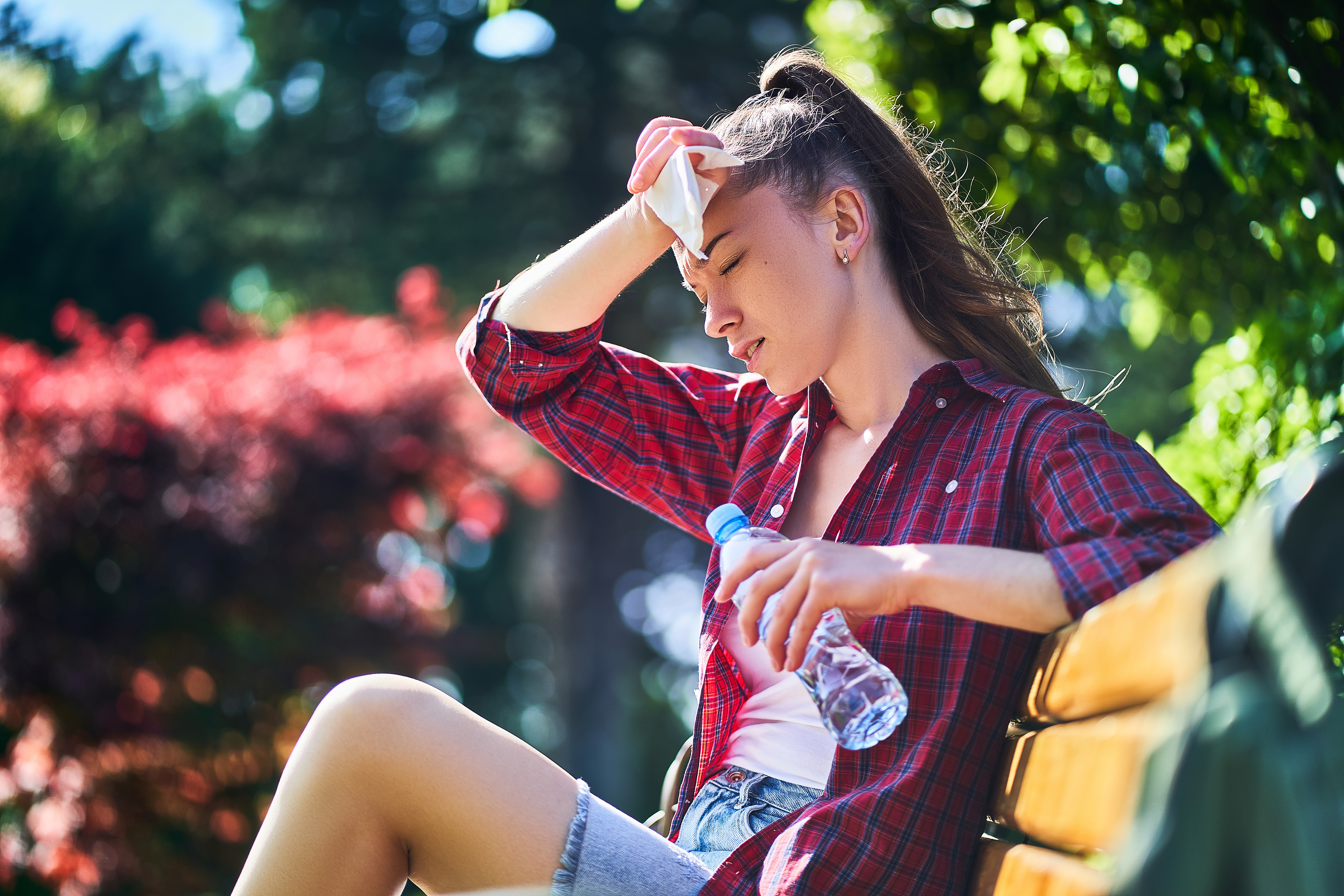 Tired sweating woman wipes her forehead with a napkin and holds cold water bottle in a park in summer hot weather